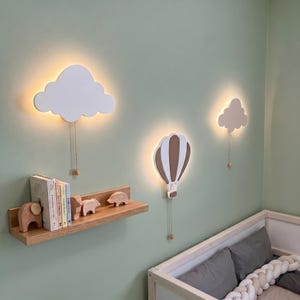 May include: Two white cloud-shaped wall lamps and one brown and white hot air balloon-shaped wall lamp, each featuring a pull chain. A wooden shelf below holds several books and three small wooden animal figurines, including an elephant.