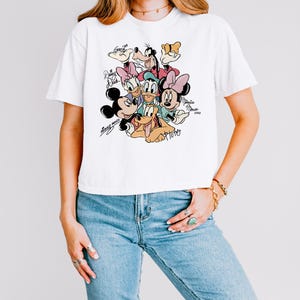 May include: White t-shirt featuring a colorful graphic of Disney characters including Mickey Mouse, Minnie Mouse, Donald Duck, Daisy Duck, Goofy, and Pluto. The shirt has the characters' names in a handwritten style. The shirt is worn by a person.