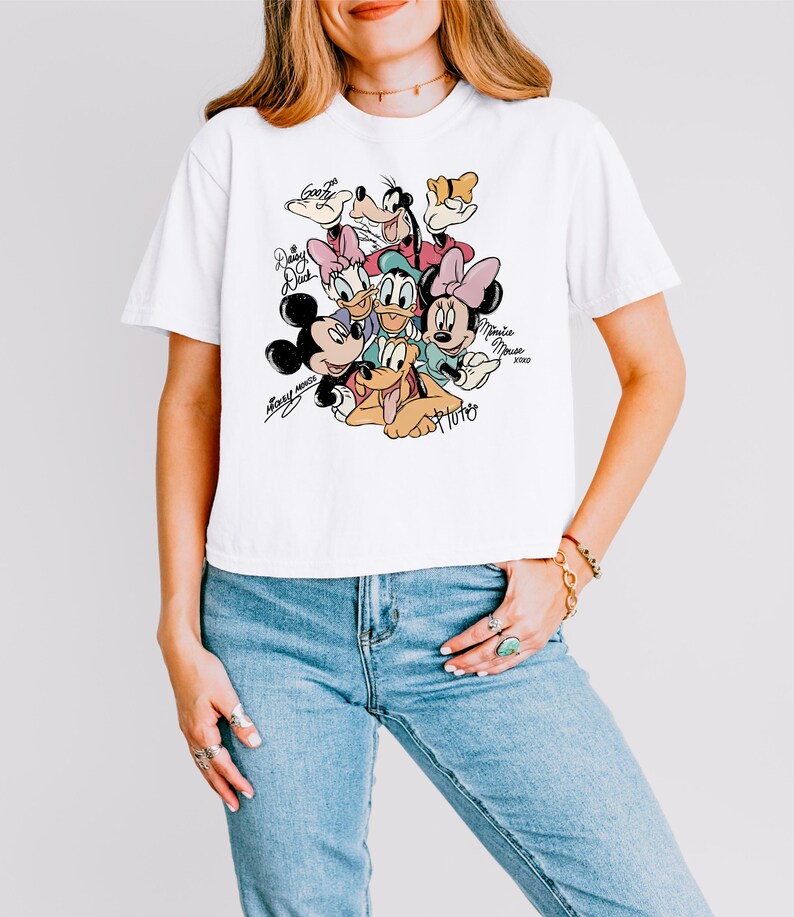 May include: White t-shirt featuring a colorful graphic of Disney characters including Mickey Mouse, Minnie Mouse, Donald Duck, Daisy Duck, Goofy, and Pluto. The shirt has the characters' names in a handwritten style. The shirt is worn by a person.
