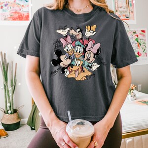 May include: Dark grey t-shirt featuring a colorful graphic of Disney characters including Mickey Mouse, Minnie Mouse, Donald Duck, Daisy Duck, Goofy, and Pluto. The shirt has the names of the characters in a script font.