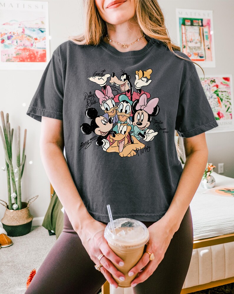 May include: Dark grey t-shirt featuring a colorful graphic of Disney characters including Mickey Mouse, Minnie Mouse, Donald Duck, Daisy Duck, Goofy, and Pluto. The shirt has the names of the characters in a script font.