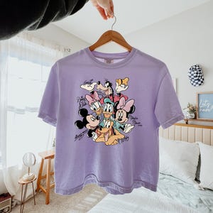 May include: A light purple t-shirt featuring a graphic of Disney characters including Mickey Mouse, Minnie Mouse, Donald Duck, Daisy Duck, Goofy, and Pluto. The shirt is hanging on a wooden hanger.