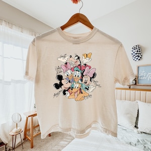 May include: Cream-colored t-shirt featuring a colorful graphic of Disney characters including Mickey Mouse, Minnie Mouse, Donald Duck, Daisy Duck, Goofy, and Pluto. The names of the characters are written in a script font. The shirt is hanging on a wooden hanger.