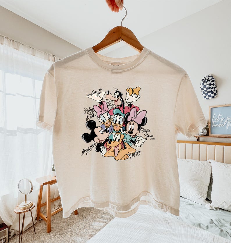 May include: Cream-colored t-shirt featuring a colorful graphic of Disney characters including Mickey Mouse, Minnie Mouse, Donald Duck, Daisy Duck, Goofy, and Pluto. The names of the characters are written in a script font. The shirt is hanging on a wooden hanger.