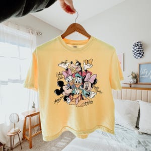 May include: Yellow t-shirt featuring a graphic of Disney characters including Mickey Mouse, Minnie Mouse, Donald Duck, Daisy Duck, Goofy, and Pluto. The characters are surrounded by their signatures. The shirt is hanging on a wooden hanger.