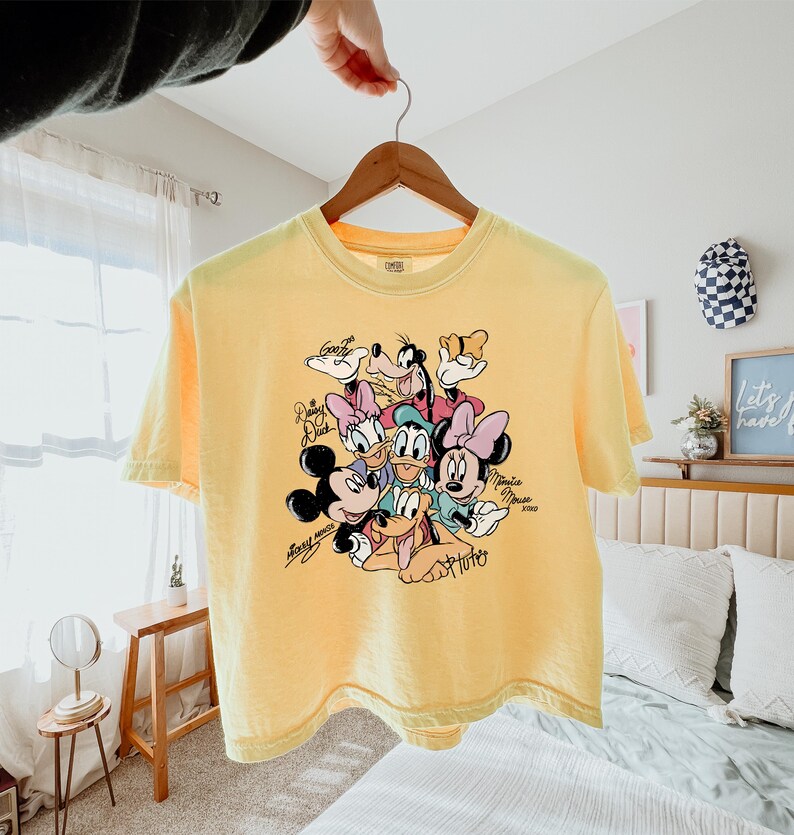 May include: Yellow t-shirt featuring a graphic of Disney characters including Mickey Mouse, Minnie Mouse, Donald Duck, Daisy Duck, Goofy, and Pluto. The characters are surrounded by their signatures. The shirt is hanging on a wooden hanger.