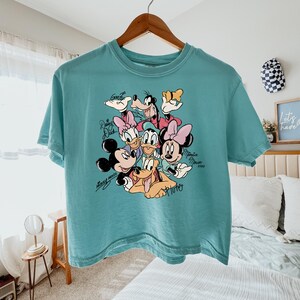 May include: Teal-colored t-shirt featuring a graphic of Disney characters including Mickey Mouse, Minnie Mouse, Donald Duck, Daisy Duck, Goofy, and Pluto. The shirt is hanging on a wooden hanger.