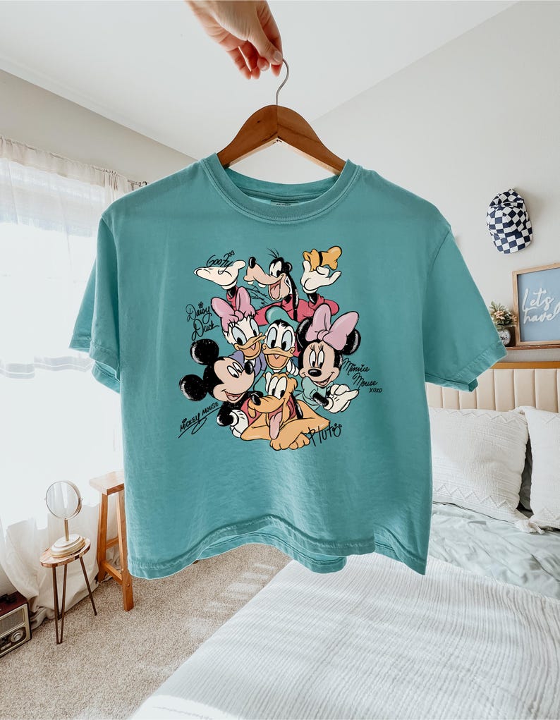 May include: Teal-colored t-shirt featuring a graphic of Disney characters including Mickey Mouse, Minnie Mouse, Donald Duck, Daisy Duck, Goofy, and Pluto. The shirt is hanging on a wooden hanger.