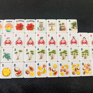 Puede incluir: Un conjunto de fichas de Mahjong blancas con ilustraciones coloridas. Las fichas presentan im&aacute;genes de flores, cangrejos, palmeras y diversas frutas. Algunas fichas tienen las palabras "JOKER", "FLOWER", "SOAP", "GREEN" y "RED".