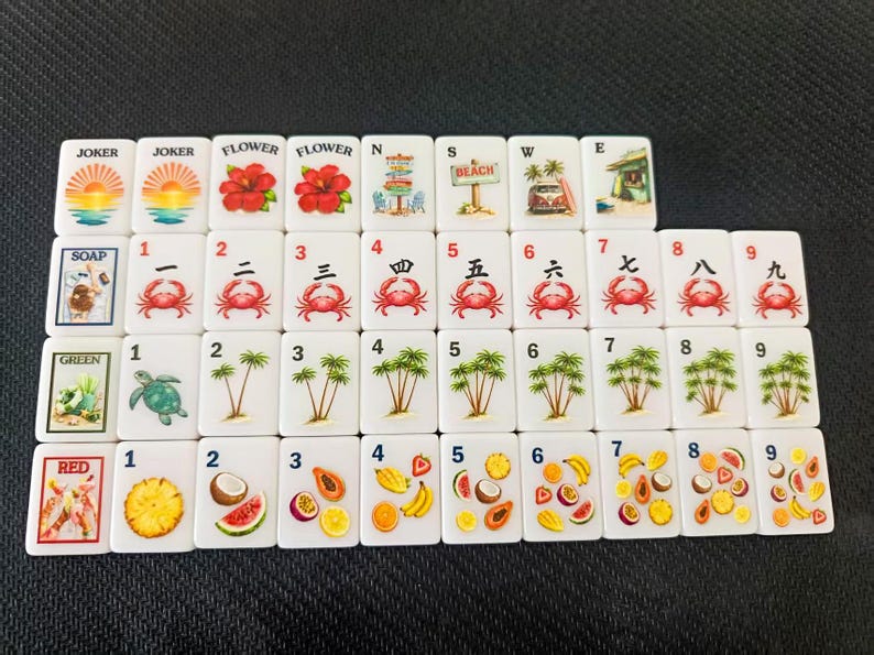 Puede incluir: Un conjunto de fichas de Mahjong blancas con ilustraciones coloridas. Las fichas presentan im&aacute;genes de flores, cangrejos, palmeras y diversas frutas. Algunas fichas tienen las palabras "JOKER", "FLOWER", "SOAP", "GREEN" y "RED".