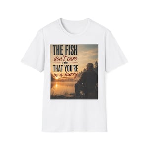 Könnte beinhalten: Ein weißes Kurzarm-T-Shirt mit einem Grafikdruck, der eine Person beim Angeln an einem See bei Sonnenuntergang zeigt. Der Text lautet: THE FISH don't care THAT YOU'RE in a hurry. Das Design enthält die Silhouette einer Person mit einer Angelrute.