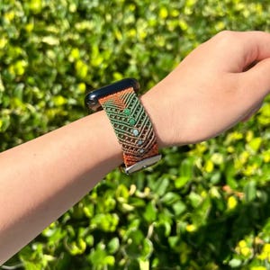 Puede incluir: Un Apple Watch con una correa tejida en tonos naranja, verde y marrón. La correa presenta un patrón de chevrón con pequeñas cuentas de color azul claro y verde. La caja del reloj es negra y la correa se asegura con un cierre plateado.