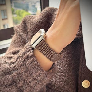 以下が含まれることがあります： シルバーカラーのスマートウォッチと、ダークブラウンの編み込みコードバンド。バンドは一連の垂直な結び目で構成されており、デザイン全体に小さな金属製のビーズが均等に配置され、手首に時計を固定しています。