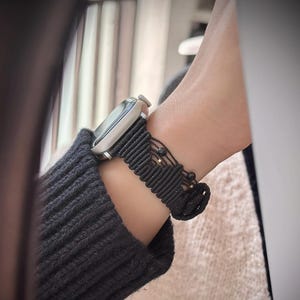Może przedstawiać: Srebrny smartwatch z czarnym paskiem z makramy jest noszony na nadgarstku. Pasek ma tkany wzór z małymi złotymi koralikami. Osoba nosi czarny sweter. Zegarek to nowoczesny dodatek.