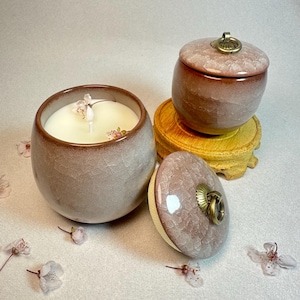 Può includere: Una candela in ceramica in un contenitore rotondo grigio-marrone con una candela bianca e un accento floreale. Un barattolo abbinato con coperchio e manico in ottone poggia su un supporto di legno. Il coperchio è rimosso, rivelando un interno color crema.