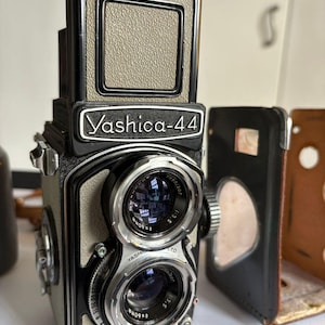 May include: A vintage Yashica-44 twin-lens reflex camera. The camera features a black and silver body with dual lenses and a top-mounted viewfinder. The Yashica-44 logo is prominently displayed. A leather case is visible to the right.