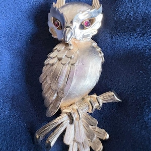 Op de afbeelding: Goudkleurige uilbroche met getextureerde veren en takzit. De uil heeft rode edelsteenogen en zit op een getextureerde tak. Een accessoire in vintage-stijl.