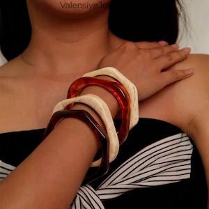 May include: A collection of geometric bangles in shades of brown, cream, and amber. The square-shaped bracelets are stacked on a wrist, showcasing their varying colors and textures. The jewelry is worn against a black and white striped top.