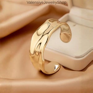 May include: A gold-colored cuff bracelet with a textured, wavy design. The open-ended bracelet has a smooth, polished finish and is displayed on a satin surface. The jewelry piece is presented in a white jewelry box.