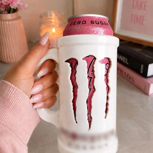 May include: A white ceramic mug designed as a holder for a beverage can. The mug features a large cutout on the front revealing the pink and silver surface of a Monster Energy Zero Sugar can. The mug has a sturdy handle and a cylindrical shape.