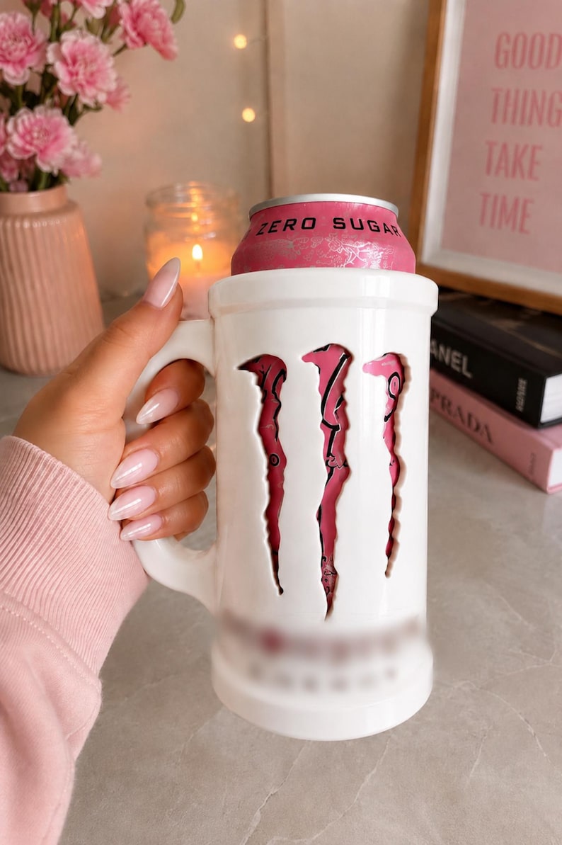 May include: A white ceramic mug designed as a holder for a beverage can. The mug features a large cutout on the front revealing the pink and silver surface of a Monster Energy Zero Sugar can. The mug has a sturdy handle and a cylindrical shape.