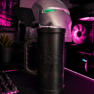 May include: A tall, cylindrical black mug with a textured, glittery finish and a sturdy side handle. The mug features a subtle embossed logo on the front surface. It is positioned upright, showcasing a sleek, dark design that contrasts with the surrounding environment.