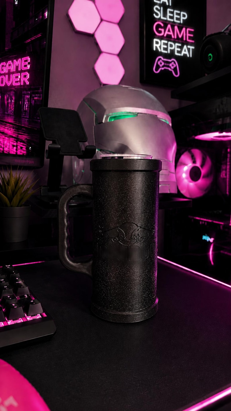 May include: A tall, cylindrical black mug with a textured, glittery finish and a sturdy side handle. The mug features a subtle embossed logo on the front surface. It is positioned upright, showcasing a sleek, dark design that contrasts with the surrounding environment.