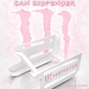 May include: A white plastic can dispenser featuring the Monster logo in pink. The unit is designed to hold 6 cans, with text stating it fits 500ml and 16 oz cans. The structure consists of two parallel side panels connected by horizontal support bars.