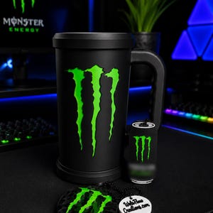 May include: A matte black cylindrical mug featuring a large neon green Monster Energy logo on the front. Beside it sits a small black canister with a matching green logo and a circular black keychain with the same logo and a white tag reading WebFlams Creations.com.