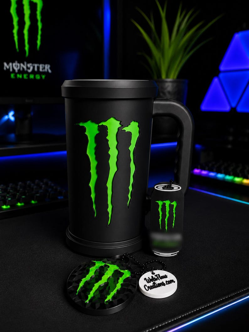 May include: A matte black cylindrical mug featuring a large neon green Monster Energy logo on the front. Beside it sits a small black canister with a matching green logo and a circular black keychain with the same logo and a white tag reading WebFlams Creations.com.