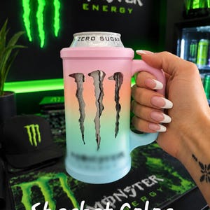 May include: A cylindrical beverage can cooler with a handle, featuring a pastel gradient finish transitioning from pink to orange to light blue. The cooler has a cutout of the Monster Energy logo, revealing a silver Zero Sugar can inside.