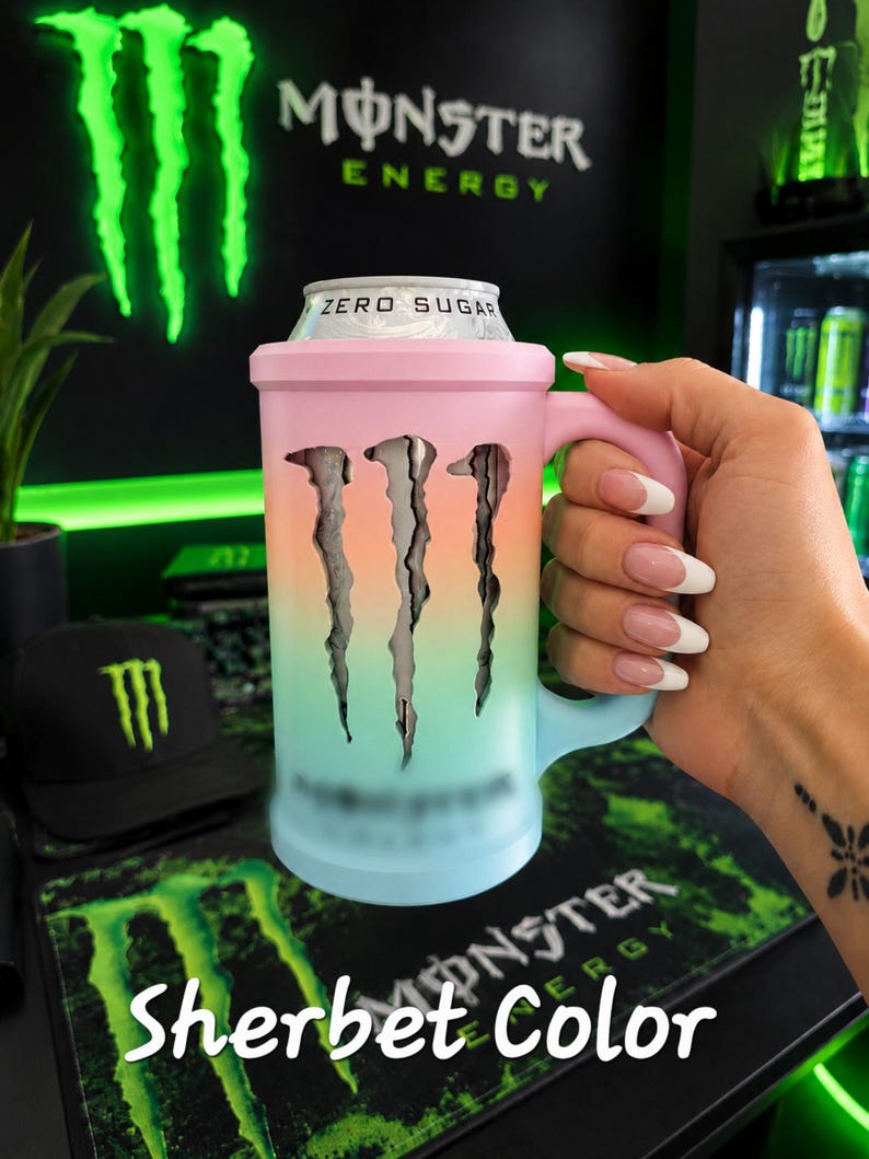 May include: A cylindrical beverage can cooler with a handle, featuring a pastel gradient finish transitioning from pink to orange to light blue. The cooler has a cutout of the Monster Energy logo, revealing a silver Zero Sugar can inside.