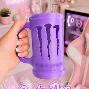 May include: A purple cylindrical mug featuring a cutout of the Monster Energy logo on the front. The mug has a sturdy handle on the side and a smooth, matte finish. The design includes a recessed area where the iconic claw mark graphic is visible.
