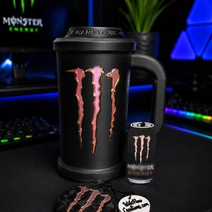 May include: A black mug featuring the Monster Energy logo in a metallic pink and orange gradient, with the text L-CARNITINE embossed on the lid. Included is a small Monster Energy can-shaped keychain and a round black medallion with the logo.