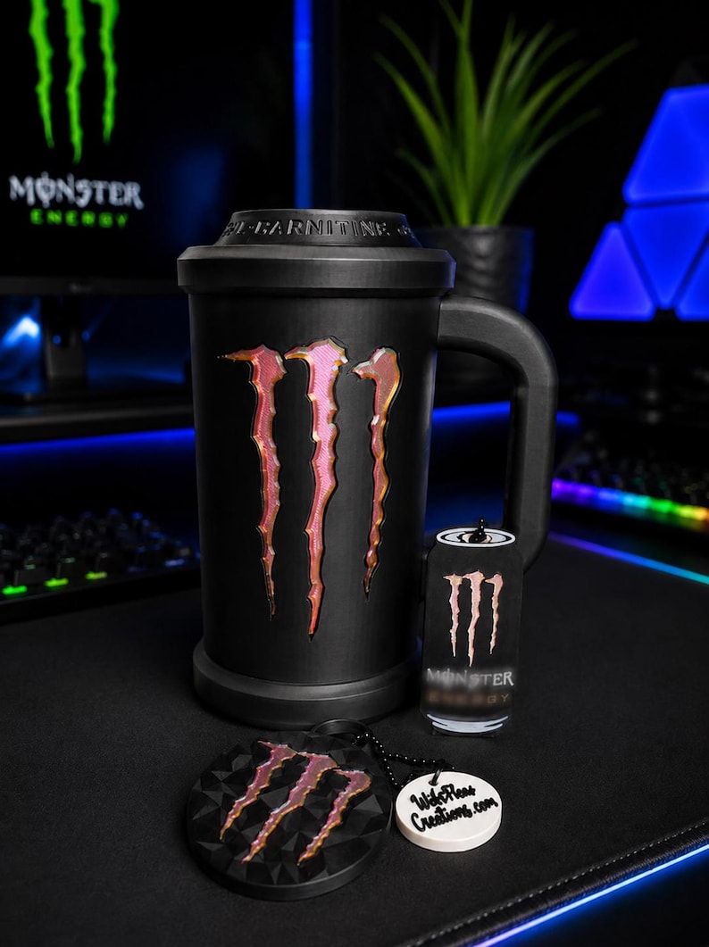 May include: A black mug featuring the Monster Energy logo in a metallic pink and orange gradient, with the text L-CARNITINE embossed on the lid. Included is a small Monster Energy can-shaped keychain and a round black medallion with the logo.