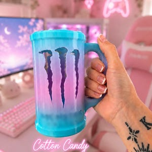 May include: A cylindrical mug with a handle, featuring a blue-to-pink gradient finish. The side displays a cutout of the Monster Energy logo, revealing the interior. The text Cotton Candy is printed in white script at the bottom of the mug.
