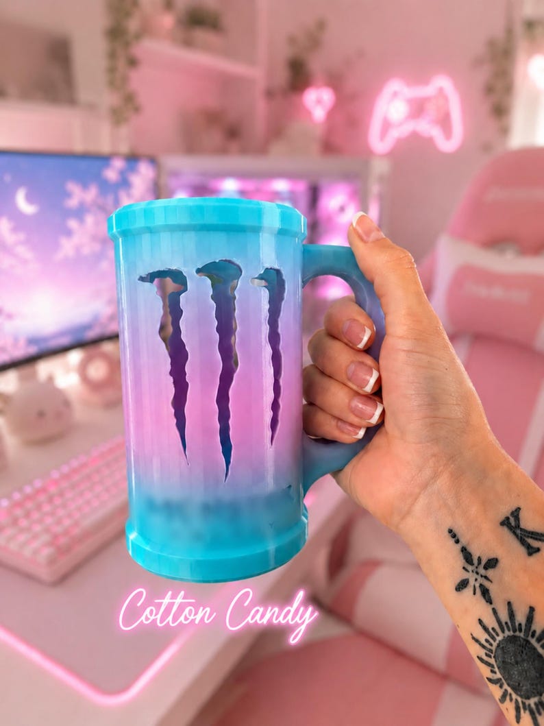 May include: A cylindrical mug with a handle, featuring a blue-to-pink gradient finish. The side displays a cutout of the Monster Energy logo, revealing the interior. The text Cotton Candy is printed in white script at the bottom of the mug.