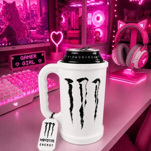 Op de afbeelding: Witte Monster Energy-blikjeskoeler met een handvat en het Monster Energy-logo. De koeler bevat een zwart blikje met het woord "TAURINE". Een label met het Monster Energy-logo hangt aan het handvat. De achtergrond omvat een roze gaming-opstelling.