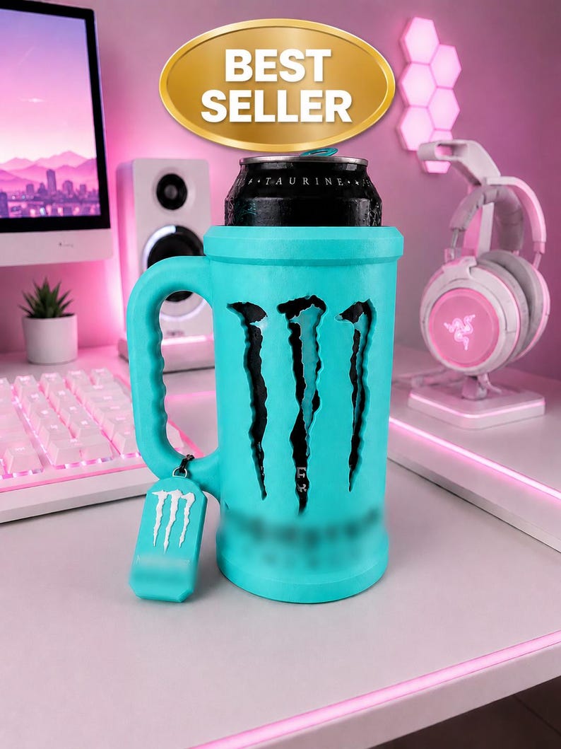 May include: A bright turquoise beverage can holder shaped like a mug with a handle. The holder features a cutout of the Monster Energy logo, revealing a black can inside. A small matching turquoise tag with the white logo hangs from the handle.