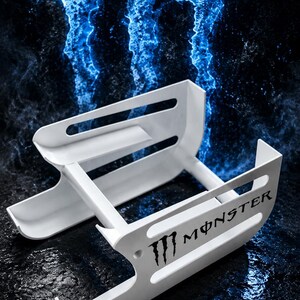 May include: A white plastic can dispenser featuring the Monster logo and text on the side. The structure consists of two parallel rails with horizontal slots, designed to hold beverage cans in a horizontal position for organized storage.
