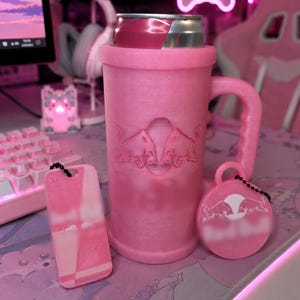 May include: A pink glittery can cooler mug with a handle, featuring an embossed Red Bull logo. Accompanied by a rectangular pink keychain and a circular pink keychain, both displaying the Red Bull logo and attached with black beaded chains.