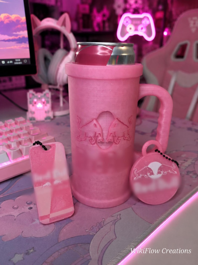 May include: A pink glittery can cooler mug with a handle, featuring an embossed Red Bull logo. Accompanied by a rectangular pink keychain and a circular pink keychain, both displaying the Red Bull logo and attached with black beaded chains.