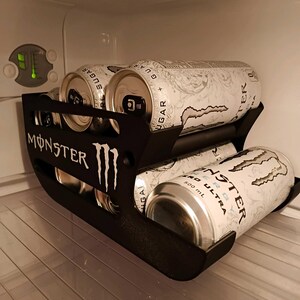 May include: A black plastic beverage can organizer rack featuring the Monster Energy logo on the side. The rack holds multiple white Monster Energy Zero Ultra cans, each labeled 500 mL, arranged in two tiers within a refrigerator interior.