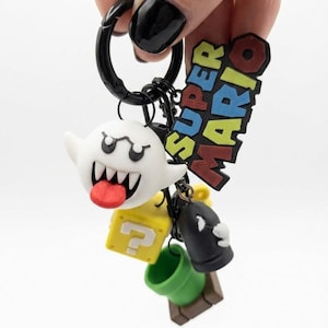 May include: A Super Mario keyring featuring a white Boo ghost with a red tongue, a yellow question mark block, a black Bullet Bill, and a green pipe. The keyring also includes a Super Mario logo.