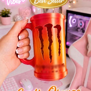 May include: A cylindrical orange and red gradient mug featuring a vertical cutout design of three claw marks on the front. The mug has a sturdy handle on the side and a smooth, solid base. The surface displays a vibrant, warm color transition.