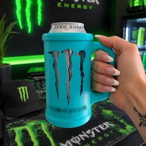 May include: A bright turquoise beverage can cooler featuring a handle and a cutout design of the Monster Energy logo. The cooler holds a silver can labeled Zero Sugar, with the iconic claw mark emblem visible through the turquoise casing.