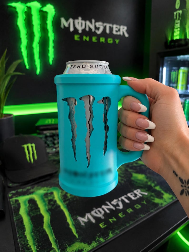 May include: A bright turquoise beverage can cooler featuring a handle and a cutout design of the Monster Energy logo. The cooler holds a silver can labeled Zero Sugar, with the iconic claw mark emblem visible through the turquoise casing.