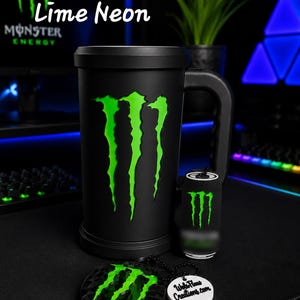 May include: A matte black cylindrical mug featuring a large neon green Monster Energy claw logo, accompanied by a small matching cylindrical keychain and a circular black and green logo keychain with a white tag reading WebFlaws Creations.com.