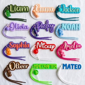 May include: A collection of colorful name tags with various names like Liam, Emma, and Jackson. Each tag features a unique color scheme and a small charm, such as a gummy bear or a lollipop, attached to a braided cord.