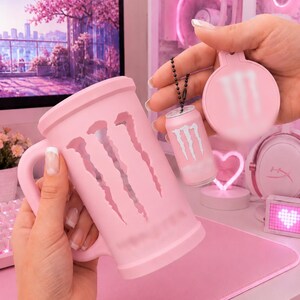 May include: A light pink mug featuring a large cutout of the Monster Energy logo on the side. Accompanying the mug is a small, matching light pink miniature can keychain and a circular light pink compact mirror, both displaying the Monster logo.